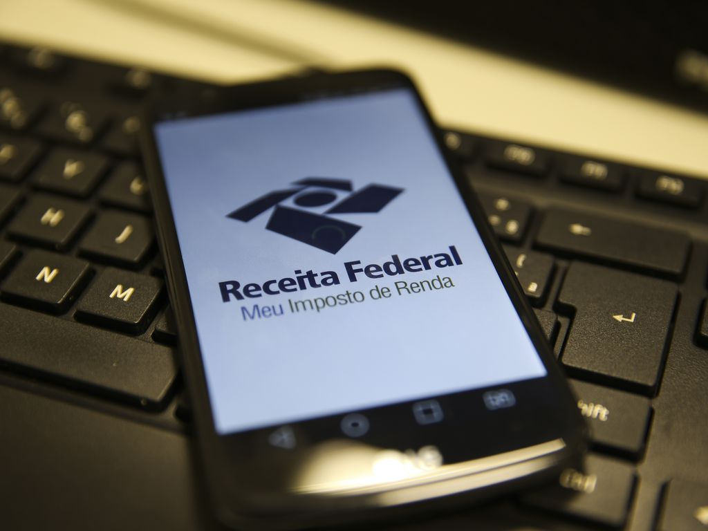 Celular exibindo o aplicativo da Receita Federal com a mensagem 'Meu Imposto de Renda' sobre teclado de computador.