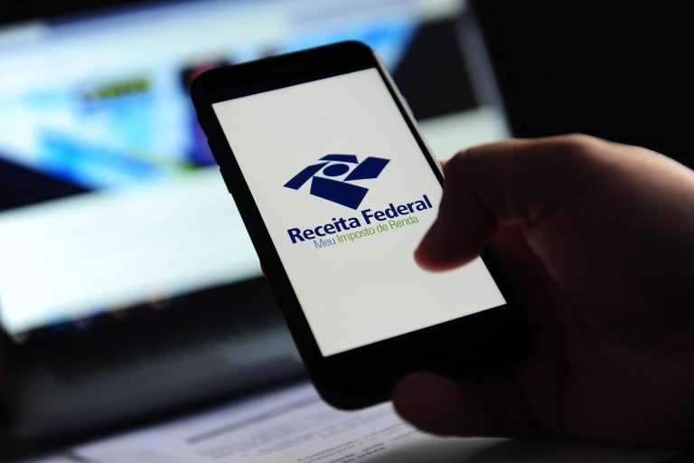 Pessoa segurando celular com a tela do aplicativo da Receita Federal, mostrando o logotipo e a mensagem 'Meu Imposto de Renda'.